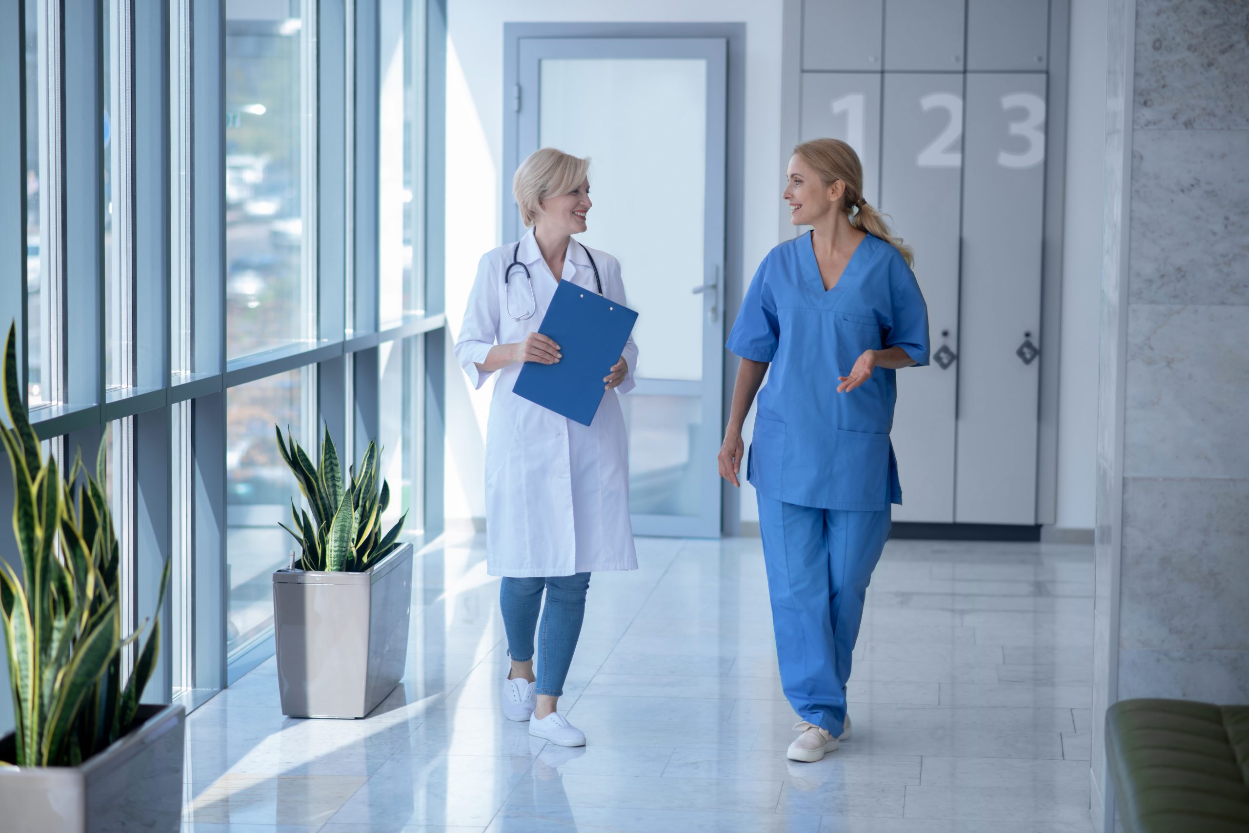 Two female doctors having friendly conversation, walking along hospital hallway